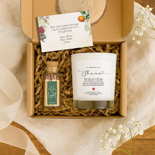 A white soy candle with the message “Your life was a blessing…” and personalised name on a transparent label, placed on a wood slice next to a match bottle labeled “Let There Be Light”, with a kraft gift box in the background. A sentimental sympathy or memorial gift.