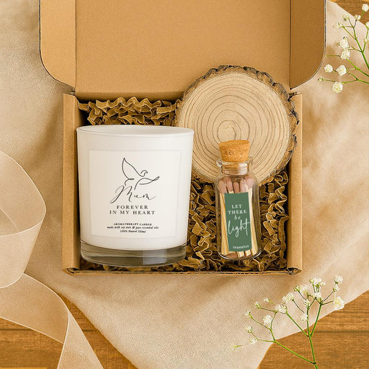 A white soy wax candle with a dove illustration and the words “Mum – Forever in My Heart” on a transparent label, displayed on a wooden slice next to a glass match bottle labeled “Let There Be Light”, with a kraft gift box in the background. A comforting sympathy or memorial gift for the loss of a mother.