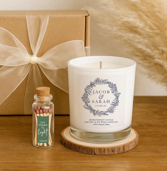 A white soy candle with a delicate grey botanical wreath and the names that can be personalised along with a wedding date, placed on a wooden slice in a kraft gift box alongside a match bottle labeled “Let There Be Light” and a floral gift note. A personalised wedding or anniversary gift set.