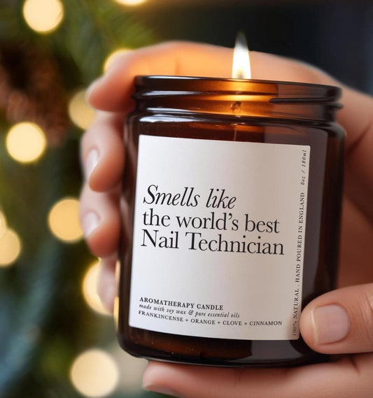 A hand holding a brown glass aromatherapy candle with a white minimalist label reading “Smells like the world’s best Nail Technician.” The candle is lit, creating a cosy, glowing atmosphere — a perfect gift for nail techs or salon professionals.