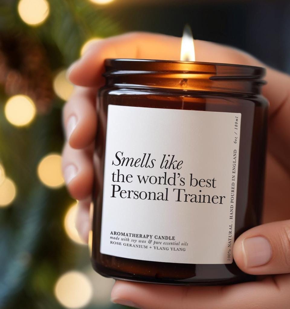 A hand holding a brown glass aromatherapy candle with a minimalist white label reading “Smells like the world’s best Personal Trainer.” The candle is lit, creating a warm glow — a thoughtful gift for a personal trainer, fitness coach, or gym instructor.