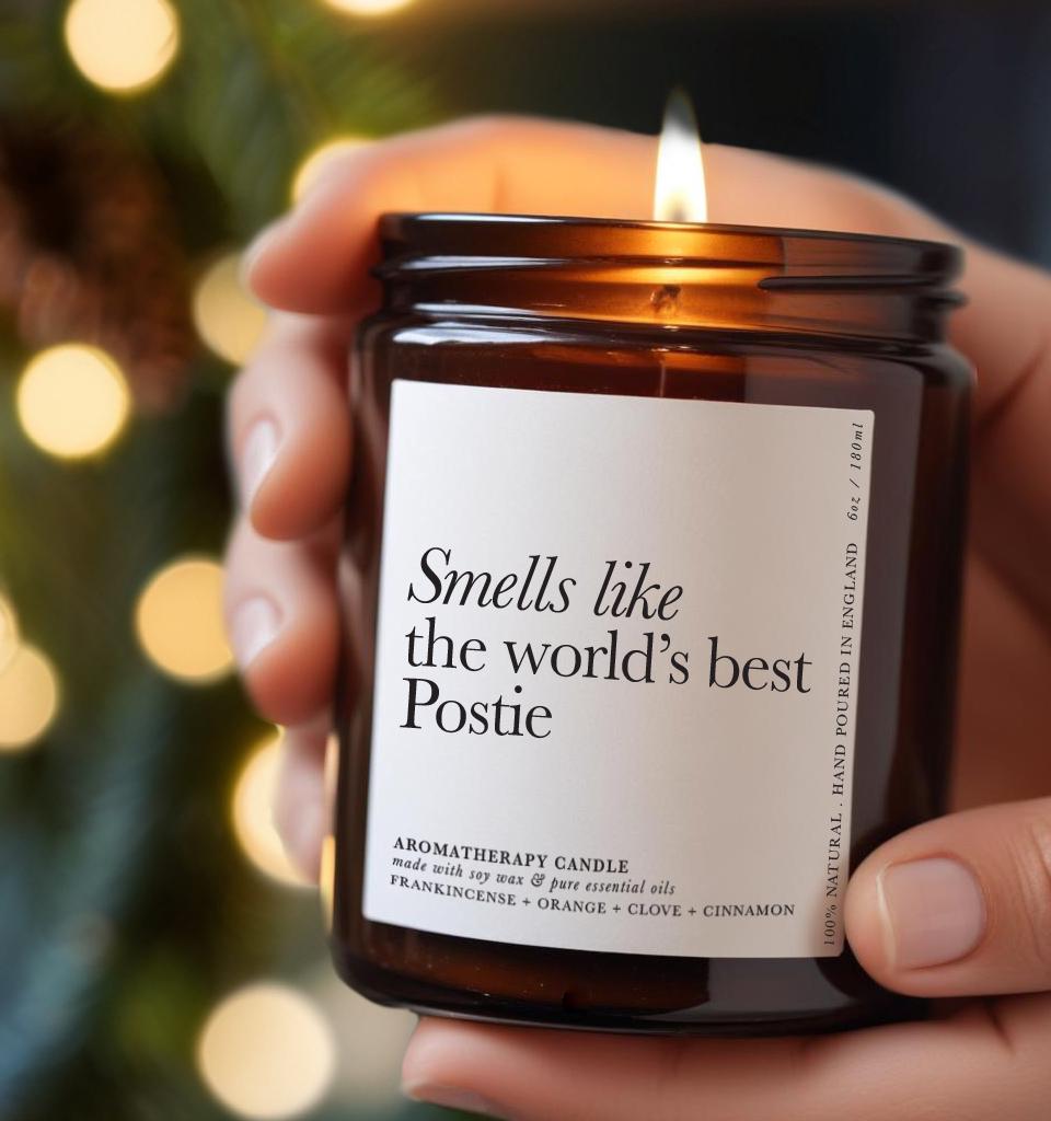 Amber glass candle with a white label reading “Smells like the world’s best Postie,” held in hands with warm festive lights in the background — ideal Christmas gift for a postman or Royal Mail postie.