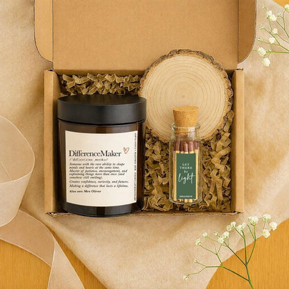 May include: A gift box with a black-lidded candle, a small glass bottle of matches, and a wooden coaster. The candle has the text "DifferenceMaker" and a definition. The match bottle reads "Let There Be Light". The box is filled with brown packing material.