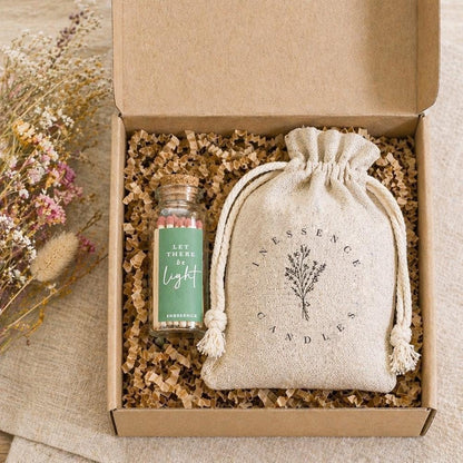 A gift box containing a small glass bottle with a label, a white cloth bag with a floral design, and some shredded paper.